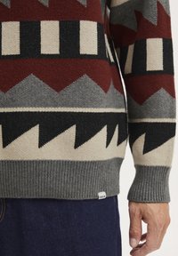 Knitted sweater featuring a geometric pattern in burgundy, gray, black, and cream, with ribbed cuffs and hem; tag labeled "JSOLID."