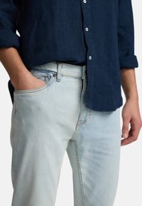 Jeans de mezclilla azul claro con un ajuste relajado, con costuras en contraste, diseño de cinco bolsillos y cierre de botón, combinados con una camisa azul oscuro.