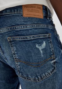 Modré džínové kalhoty s vybledlým vzhledem, viditelným šitím, zadní kapsou a hnědou koženou etiketou na pasu.