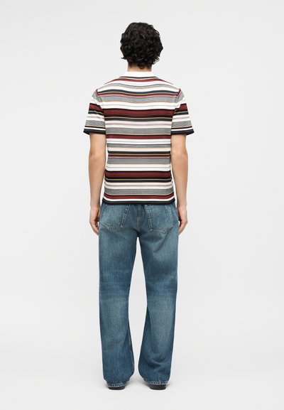 Άνδρας που στέκεται με την πλάτη στραμμένη φοράει κοντομάνικο πουκάμισο με οριζόντιες πολύχρωμες ρίγες και άνετο μπλε τζιν, μπροστά σε απλό φόντο.