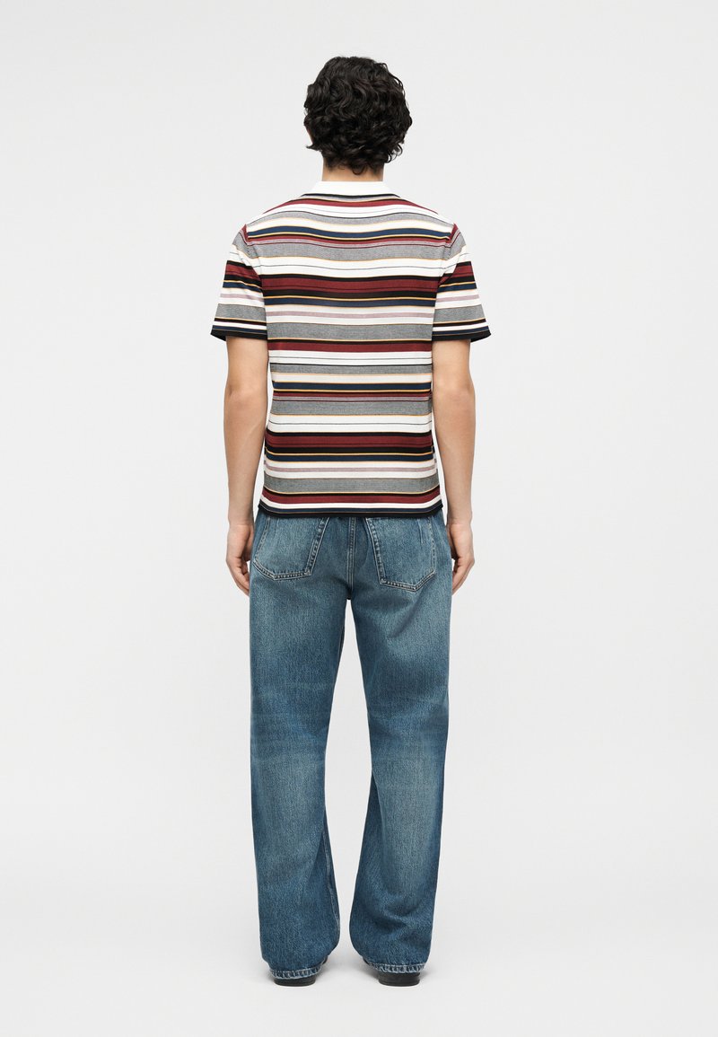 Man standing facing away wearing a multicolored horizontal striped short-sleeve shirt and loose blue jeans against a plain background.