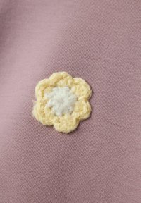 Applicazione di fiore all'uncinetto gialla e bianca su uno sfondo di tessuto lavanda, che mostra un design floreale testurizzato.