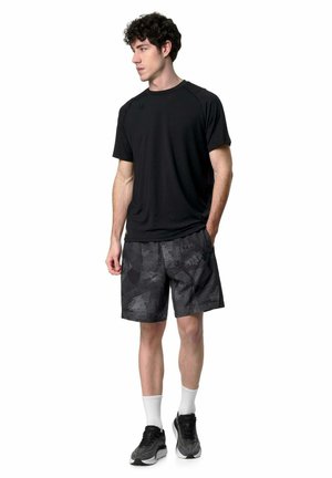 Giovane uomo in piedi che indossa una maglietta sportiva nera, pantaloncini neri con fantasia, calzini bianchi e scarpe da corsa nere, guarda verso sinistra.