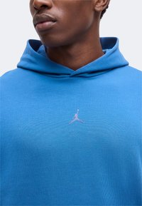 Gros plan d'une personne portant un sweat à capuche bleu avec un petit logo blanc Jumpman centré sur la poitrine.