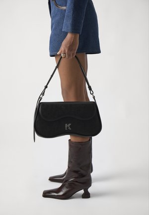 Černá kožená kabelka s zakřiveným tvarem, zdobená stříbrným logem. Rukojeť je elegantní a nastavitelná. Modelka má na sobě hnědé, lesklé kotníkové boty.