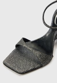 Λαμπερό μαύρο σανδάλι με ανοιχτά δάχτυλα, τετράγωνη μύτη, ένα μπροστινό λουράκι και λεπτό λουράκι στον αστράγ�αλο σε λευκό φόντο.