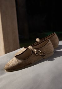 Beżowe baleriny z zamszu o zaokrąglonym nosku, z brązowym elastycznym paskiem i złoto-tonowym akcentem klamry. Płaska gumowa podeszwa.