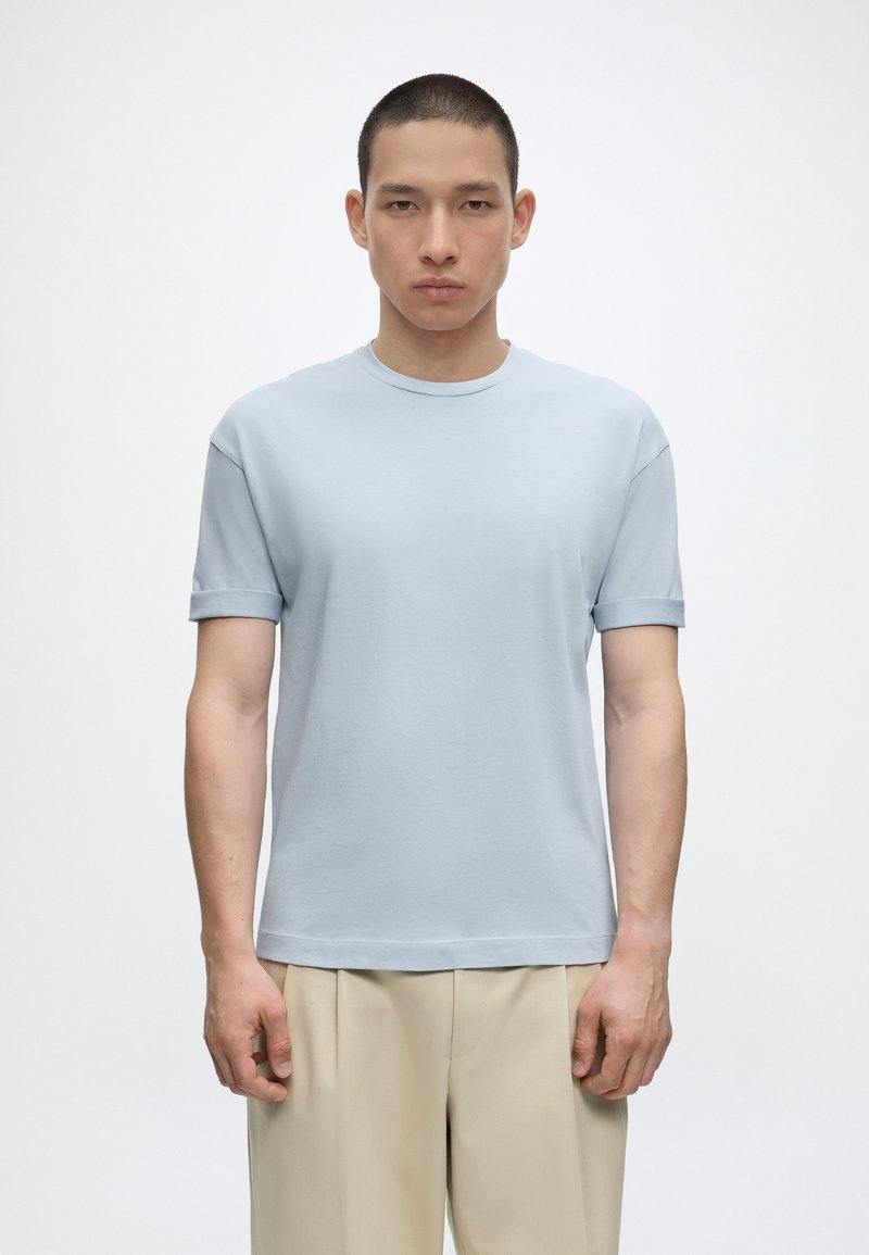 Jeune homme aux cheveux courts portant un t-shirt bleu clair à manches courtes et un pantalon beige, debout devant un fond blanc uni.