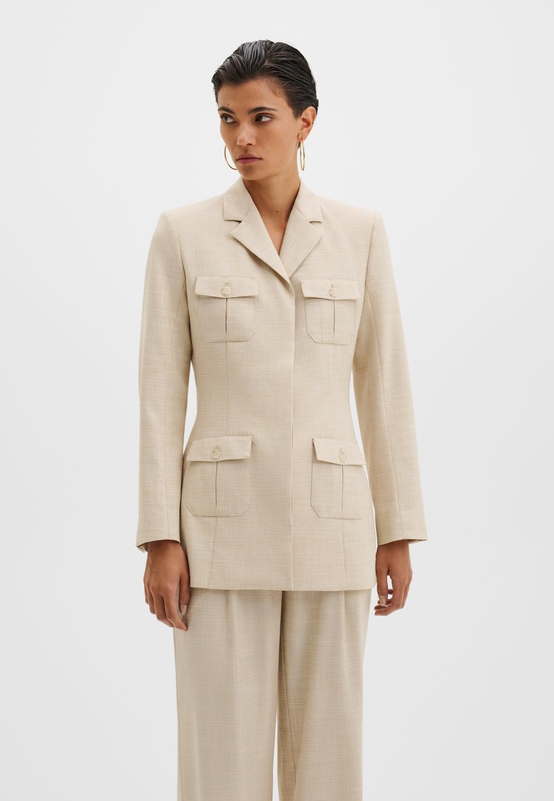 Femme portant un blazer beige ajusté avec quatre poches à boutons et un pantalon large assorti, debout devant un fond clair uni.