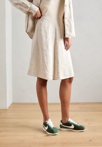Falda de lino beige con un diseño acampanado, combinada con zapatillas Nike verdes y blancas. Fondo de suelo de madera, detalles sutiles de la textura de la tela visibles.
