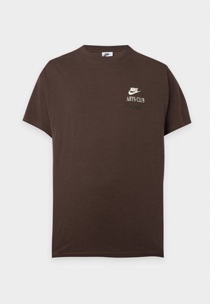 Braunes kurzärmeliges Nike T-Shirt mit weißem "Nike Arts Club"-Logo auf der linken Brust, dargestellt vor einem einfarbigen hellgrauen Hintergrund.