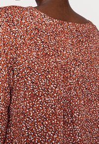 Haut orange avec un motif léopard et des accents noirs et blancs. Texture douce, encolure ronde et dos plissé.