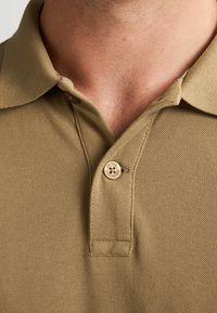 Polo shirt color khaki con un tessuto strutturato, caratterizzato da un colletto classico e un singolo bottone beige con dettagli del marchio vicino al collo.