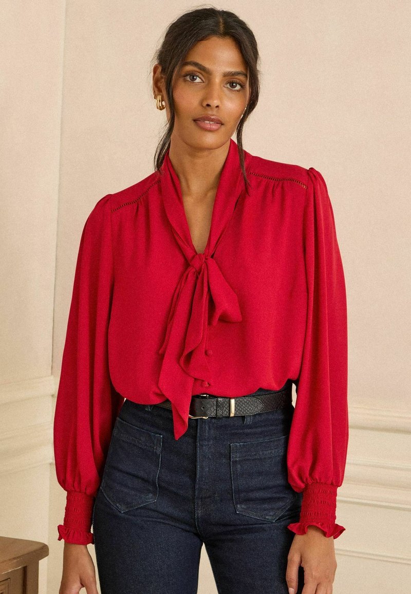 Piros blúz nyakába köthető díszítéssel és fodros elejével, hosszú puffos ujjakkal, smockolt mandzsettákkal, sötét farmernadrággal és fekete övvel viselve.