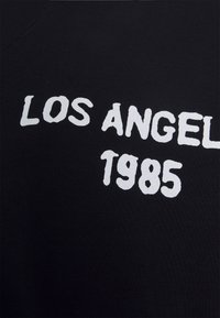 Black cotton T-shirt with white text reading "LOS ANGELES 1985." The fabric has a smooth texture and relaxed fit.