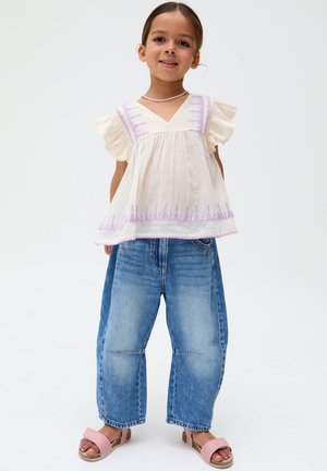 Mädchen trägt weiße Bluse mit lila Stickerei, weite blaue Jeans, pinke Sandalen und eine pinke Halskette, steht vor weißem Hintergrund.