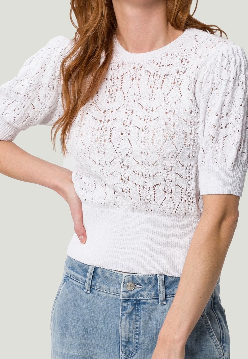 Hvid strikket sweater med korte puffærmer, der har et struktureret blonde mønster og en tætsiddende ribbet talje, parret med lyseblå denimshorts.