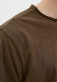 Brązowy t-shirt z okrągłym dekoltem, wykonany z lekko teksturowanej tkaniny i z surowymi krawędziami przy dekolcie i rękawach.