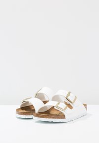 Sandalias blancas con dos correas y hebillas doradas, plantilla de corcho y suela de goma blanca. Textura suave con un diseño minimalista.