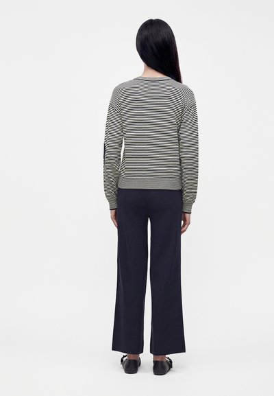 Femme aux longs cheveux noirs portant un pull à rayures noires et blanches, un pantalon large foncé et des chaussures noires, debout, vue de dos, sur fond blanc.