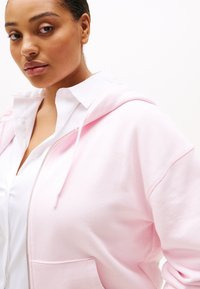 Lichtroze hoodie met een ritssluiting aan de voorkant, een trekkoordkap en voorzakken. Kenmerkt zich door subtiele branding in koninklijk reliëf op de borst.