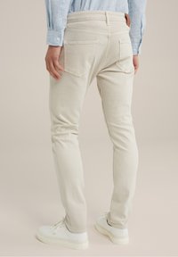 Béžové slim-fit džíny s náprsními kapsami, jemnými švy a hladkou texturou, doplněné bílými teniskami.
