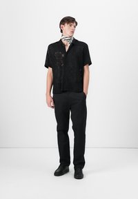 Camisa preta de manga curta com um intricado design de rendado, combinada com calças pretas e sapatos pretos. Um lenço com padrão preto e branco é usado em volta do pescoço.