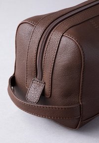 Close-up of a brown textured leather toiletry bag with a zipper and a stitched handle on a light gray surface.