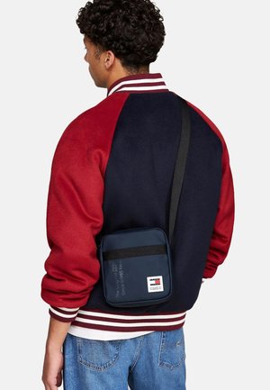 Granatowa torba na ramię z zamkiem błyskawicznym i czarnym paskiem, z naszywką z logo. Model ma na sobie czerwono-granatową kurtkę varsity w paski.