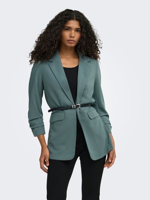Vrouw met lang krullend haar, die een teal blazer draagt, aangesnoerd met een dunne zwarte riem over een zwart topje en zwarte broek, staand tegen een effen achtergrond.