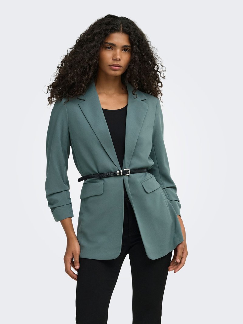 Femme aux longs cheveux bouclés portant un blazer bleu sarcelle cintré avec une fine ceinture noire sur un top noir et un pantalon noir, debout devant un fond uni.