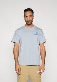 Ανοιχτό μπλε βαμβακερό T-shirt με στρογγυλή λαιμόκοψη και κοντά μανίκια, με μπλε λογότυπο στο στήθος, συνδυασμένο με μπεζ παντελόνι.