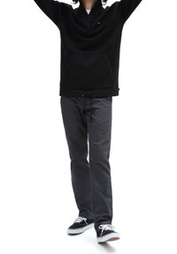 Persona che indossa una felpa nera con zip a quarto, pantaloni grigio scuro e sneakers bianco e nere, in piedi con le braccia alzate su uno sfondo bianco.