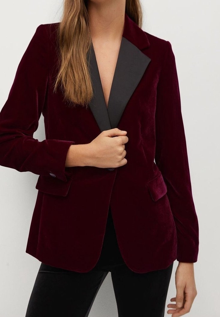 Femme portant un blazer en velours bordeaux avec des revers noirs, tenant le devant d'une main, assorti à un pantalon noir.