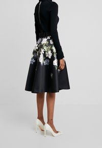 Zwarte bloemenrok met een satin afwerking, met een getailleerde taille en een uitlopende zoom. Gecombineerd met witte hoge hakken.