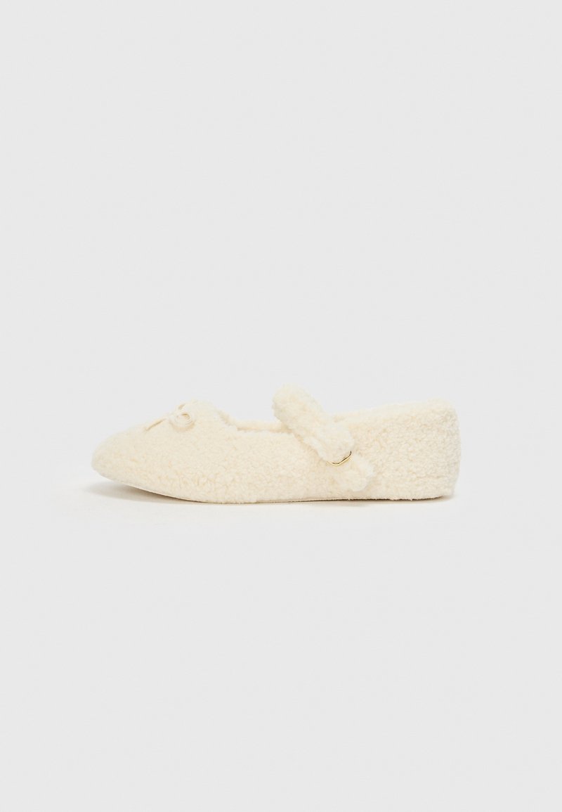 Cremefarvet hjemmesko med en fuzzy tekstur, flad design, rund tå og en dekorativ rem med en lille accent.