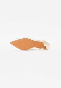 Zapato de tacón alto dorado con punta afilada, parte superior blanca tejida y suela lisa. Presenta una delicada correa alrededor del tobillo y un acabado brillante.