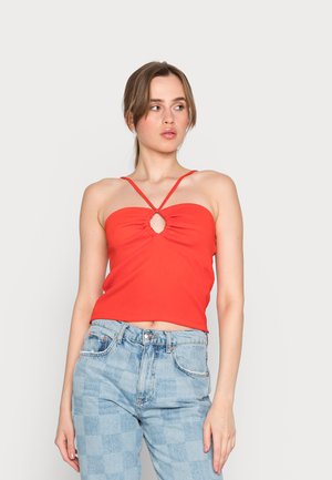 Top cropped vermelho com texturização em ribana, apresentando finas alças cruzadas e um detalhe em forma de chave no centro, combinado com jeans de cintura alta azul claro em padrão xadrez.