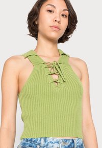 Top halter de malha canelada verde, com colarinho, frente com atacadores e design justo. A textura é suave e a cor é vibrante.