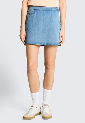 Femme portant une mini-jupe en denim bleu clair, des chaussettes blanches et des baskets beige-blanc, debout sur un fond clair.