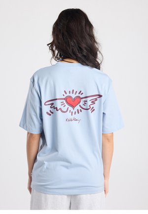 T-shirt en coton bleu clair avec un graphique de cœur rouge ailé au dos, avec "Keith Haring" imprimé dessous en bordeaux. Manches courtes.