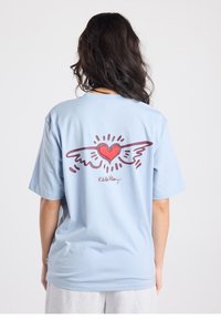 Lichtblauw katoenen T-shirt met op de achterkant een rood hart met vleugels en daaronder de tekst "Keith Haring" in bordeauxrood. Korte mouwen.