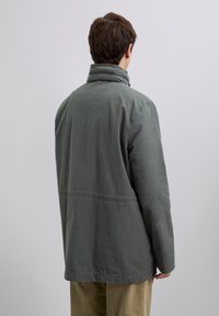 Man wearing a gray high-collar jacket and beige pants, standing facing away against a plain light gray background.