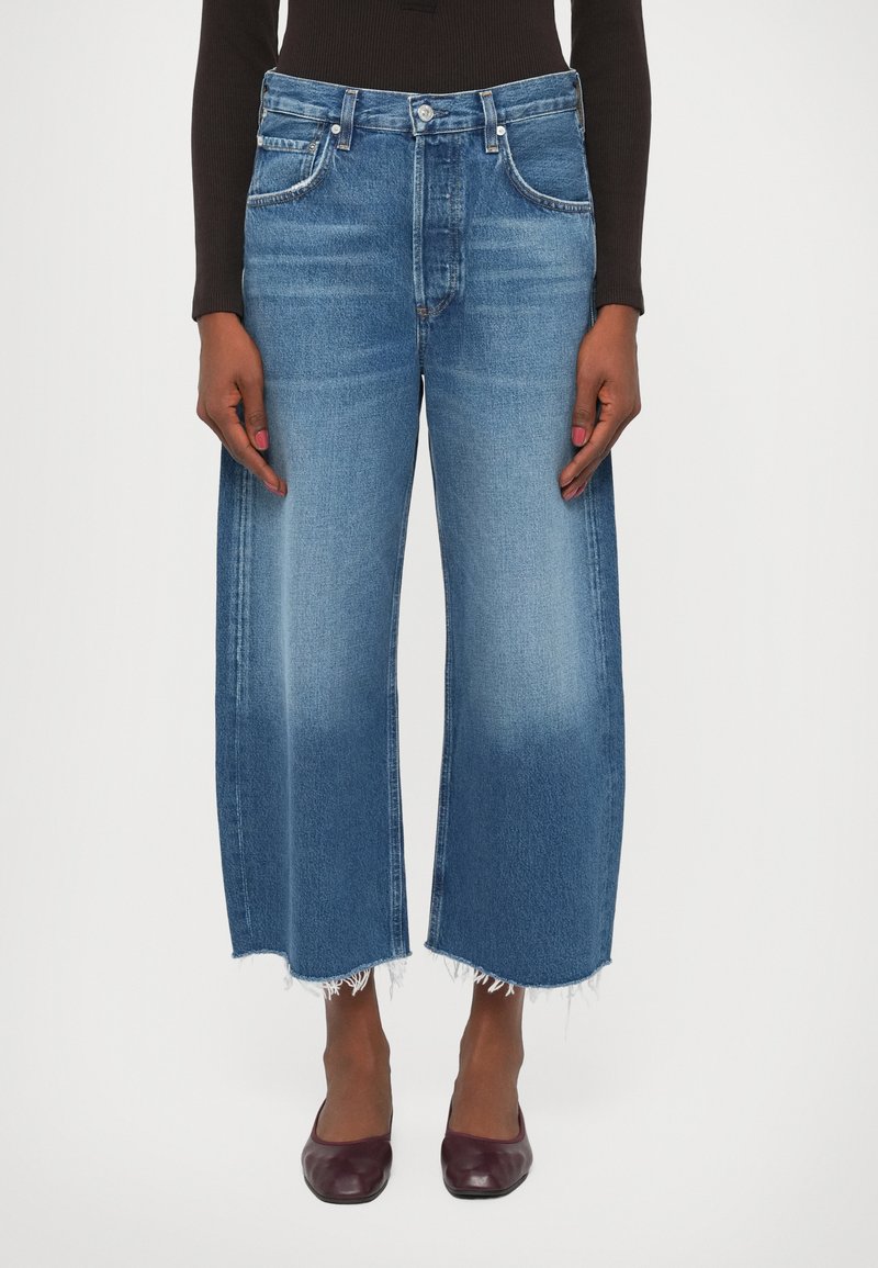 Jeans de tiro ancho en denim azul con un lavado descolorido y dobladillo sin coser. Diseño de cinco bolsillos con herrajes plateados y detalles de costura en contraste.