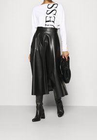 Weißes Langarmhemd mit schwarzem Grafiktext, kombiniert mit einem hochgeschnittenen schwarzen Kunstlederrock und schwarzen Stiefeletten mit Absatz. Schwarze Clutch-Tasche.