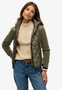 Olijfgroene gewatteerde jas met capuchon, gecombineerd met een crème kabelbrei-trui. Spijkerbroek en geweven riem, die een casual gelaagde look laat zien.