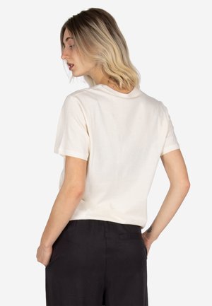 Blonde woman with shoulder-length hair wearing a white short-sleeve shirt and black pants, standing with one hand in her pocket.