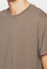 T-shirt à manches courtes et col rond en coton brun clair avec un logo embossé subtil sur la poitrine. Texture douce et lisse.