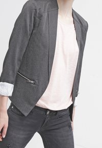 Blazer grigio sartoriale con una silhouette strutturata, tasche con cerniera e tessuto testurizzato. Indossato sopra un top rosa chiaro e jeans scuri.