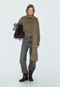 Olijfgroen sweater met een lange sjaal, gecombineerd met grijze jeans en zwarte enkellaarsjes. Het model houdt een donkere tas vast. Eenvoudig, modern ontwerp.